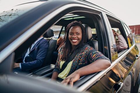 A person smiling sitting in the front seat of a car with other passengers inside